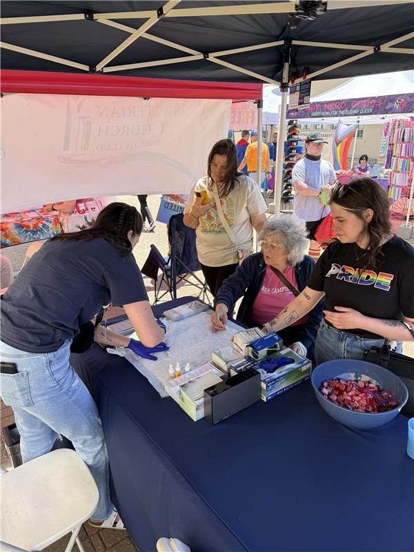Three women gathered at an ImpactLife booth under a black canopy: one volunteer leans over to prepare supplies while two others—one holding a phone—look on, with market stalls behind them.