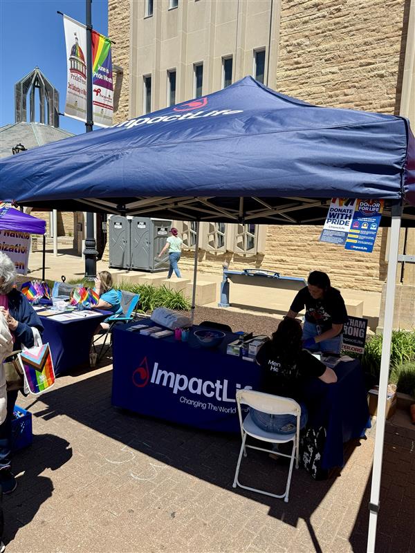 Wide shot of an ImpactLife tent and table at an outdoor event; a volunteer attends to a visitor while rainbow flags and vendor booths line the background.