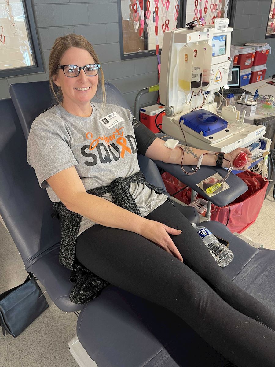 Woman wearing glasses and a 'Support Squad' T-shirt while donating blood at the ImpactLife event.