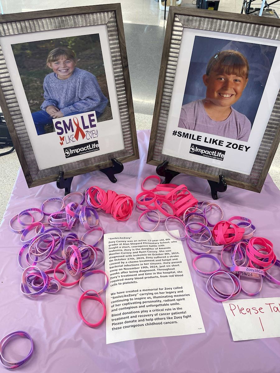 Two framed photos of Zoey labeled 'Smile Like Zoey' with pink and purple bracelets on a table at the ImpactLife event.