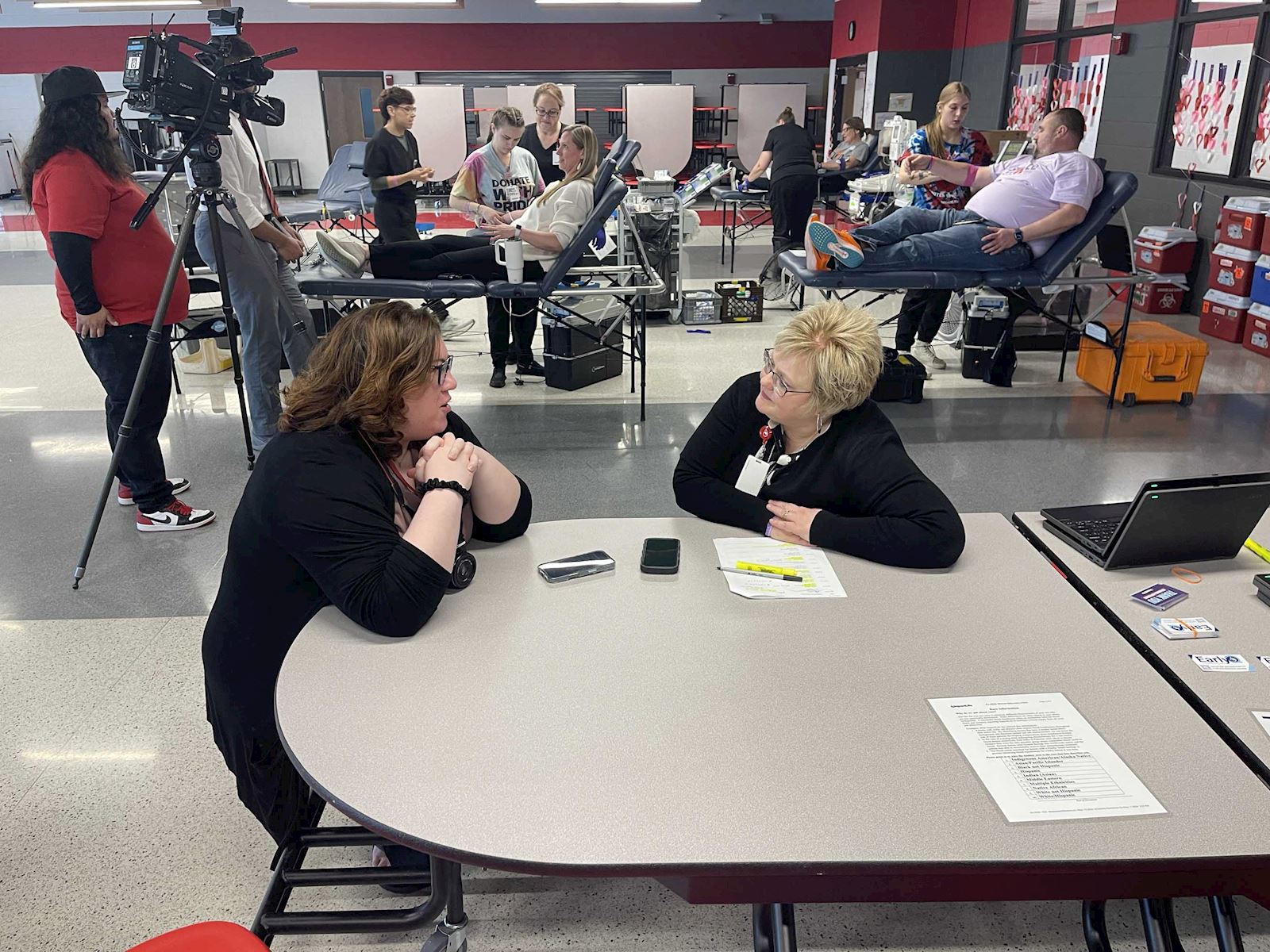 Two women talking at a table during the ImpactLife blood drive, with donors in the background.