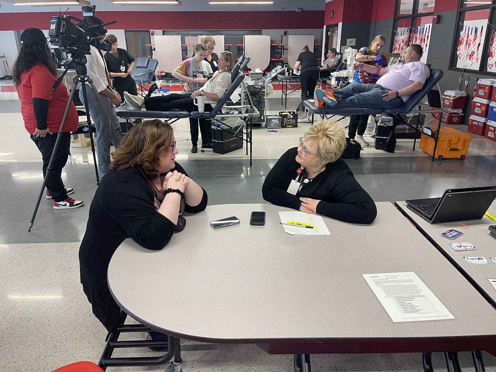Another view of the ImpactLife blood drive, showing donors and a camera crew in the background.
