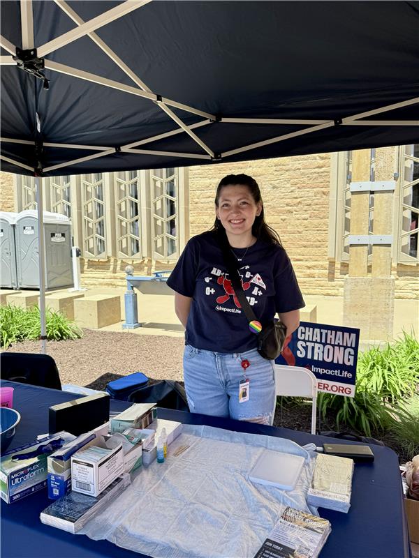 ImpactLife volunteer smiles standing behind a table laden with supplies and a ‘Chatham Strong' sign, under a black canopy at a sunny community event.